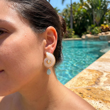 Shell drop earrings with gold trim and blue teardrop charm worn by a model outdoors near a pool