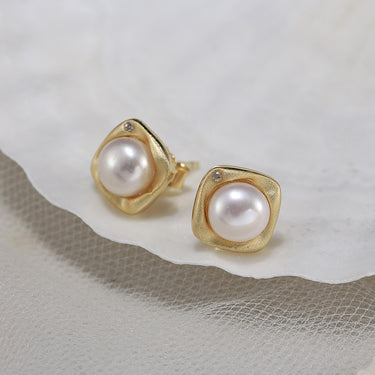 Close-up of cushion bezel gold pearl earrings on textured white fabric.