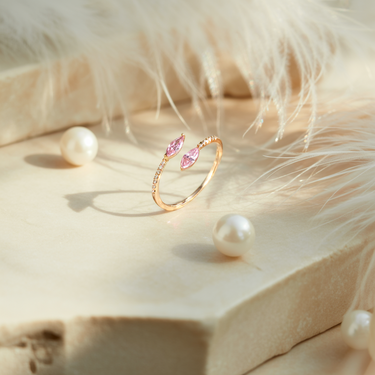Sunrise Horizon Kiss Ring with pink gemstones on a soft, beige surface with feathers