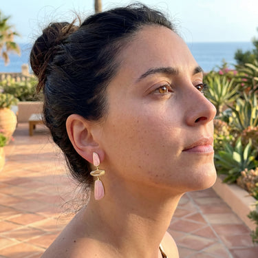 Pink and gold drop earrings with stone segments worn on a model outdoors near the ocean