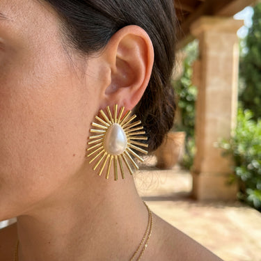 Gold sunburst stud earrings with a white teardrop pearl center, worn outdoors on a model