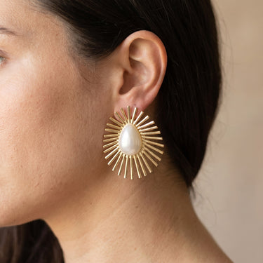 Gold-tone sunburst stud earrings with a white baroque pearl center, worn on a model
