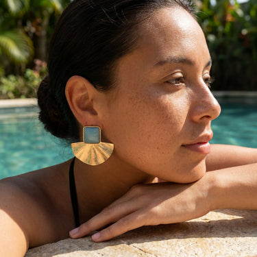 Gold brass fan earring with turquoise stone worn on a model outdoors near a pool